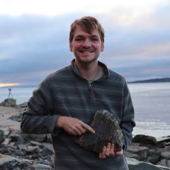 A smiling man standing on a rocky beach, holding a large stone in his hands and pointing and the different coloured stripes on it.