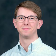 A smiling man with glasses and short light brown hair, wearing a light blue checkered shirt in front of a dark blue background. 