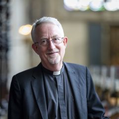 A white man wearing a clerical collar and black clothing stands in a church