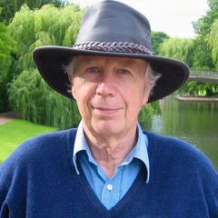 Alan Mcfarlane, a smiling man wearing a brown rimmed hat, a pale blue shirt and a dark blue jumper. He is standing in front of a green river bank