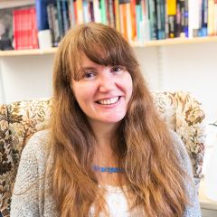 Alice Hutchings, a smiling woman with long wavy light brown hair. She is wearing a blue necklace, a white t-shirt and a grey cardigan. She is sitting in front of a book shelf