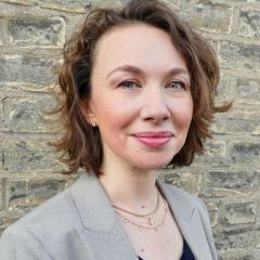 Anna Alexandrova, a smiling woman with short brown wavy hair wearing a black top and a grey blazer. She is standing in front of a brick wall.