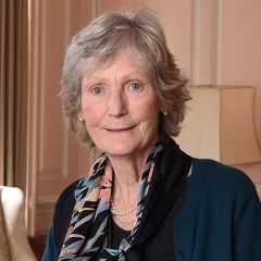 Ashley Moffett, a smiling woman with short grey hair wearing a blue and black scarf in a pale pink room