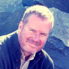 Bert Vaux, a smiling man with short fair hair and beard wearing a casual jacket. He is outside in the sun and there are rocks behind him.