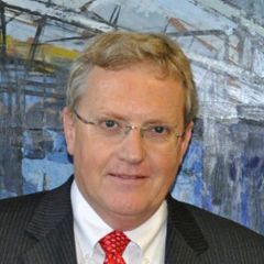 Cam Middleton, a smiling man with short fair hair and glasses, wearing a suit and tie. He is standing in front of a blue expressionist painting.