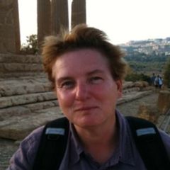 Caroline van Eck, a smiling woman with short blonde hair and a casual blue shirt. She is wearing a back pack and standing in an amphitheatre.
