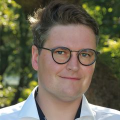 Dale Kedwards, a smiling man with short dark hair and glasses wearing a white shirt. He is outside in a green space.