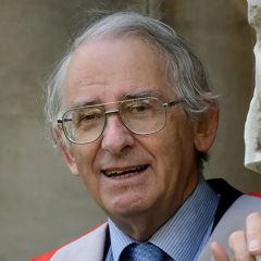 Dan McKenzie, a smiling man with grey hair and glasses. He is wearing a blue shirt and tie. He is standing outdoors in front of stone brickwork