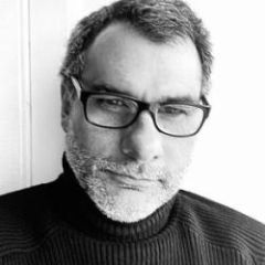 A black and white image of a man with short dark hair, rectangular glasses and a black polo neck jumper