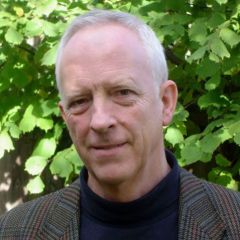 Gareth Steadman Jones, a smiling man the short grey hair wearing a navy blue polo neck and a brown tweed jacket. He is standing in front of a hedge