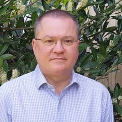 Ian James, a man with short grey hair and glasses wearing a pale shirt. He is outdoors standing against a hedge