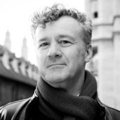 James Laidlaw, a black and white photo of a man with short curly hair wearing a scarf. He is outside in an urban environment