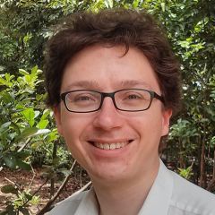 Jamie Vicary, a smiling man with brown curly hair wearing  glasses and a white shirt. He is standing outdoors and surrounded by greenery