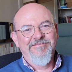 John Arnold, a smiling bald man with a short beard wearing glasses and a blue jumper