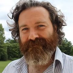 John Filling, a smiling man with curly brown hair and a beard. He is wearing a blue and white striped shirt and is outdoors in a green space.