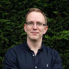 John Perry, a smiling man with shirt blond hair wearing glasses and a navy blue shirt. He is outdoors in front of a dark hedge