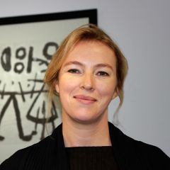 Jude Brown, a smiling woman with tied back blonde hair and a black jumper. She is standing in front of a monochrome abstract artwork.