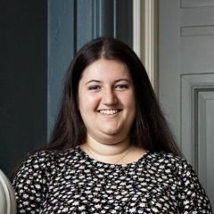 Katie Haworth, a smiling woman with long dark hair wearing a black and white floral top. She is standing in front of grey and blue painted panelling