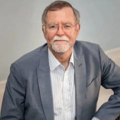 Keith Carne, a smiling man with short grey hair and beard wearing a white shirt and a grey suit