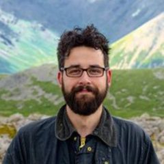 Matei Candea, a smiling man with short dark hair and a beard wearing glasses and a casual blue jacket. He is outdoors in a mountain range