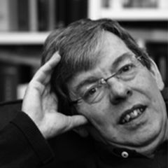 A black and white image of a man with short dark hair and glasses wearing a dark shirt. He is sitting in a relaxed pose