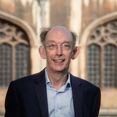Nicholas Marston, a smiling bald man wearing glasses, a pale blue shirt and a black jacket. He is standing outside the King's College Chapel