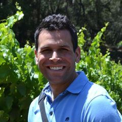 Nicholas Tosca, a smiling man with dark hair wearing a pale blue polo shirt. He is outdoors surrounded by greenery in the sun