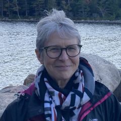 Nicolette Zeeman, a smiling woman with short grey hair and glasses wearing a geometric print scarf and a walking jacket. She is standing near a lake lined with trees