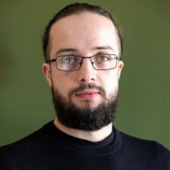 A smiling man with long, tied back, dark hair, a a beard and glasses. He is wearing a black T-shirt and is standing against a olive green background