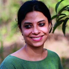 Perveez Mody, a smiling woman with dark tied back hair wearing a green top. She is outside surrounded by leaves.