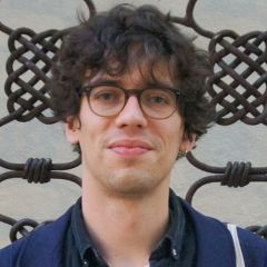 Phil Knox, a smiling man with thick dark curly hair. He is wearing glasses, a small silver earring and a casual blue jacket. He is standing in front of some decorative woven iron work.