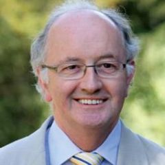 Chris Gilligan, a smiling man with short grey hair and glasses wearing a pale suit. He is standing outdoors in a green space.