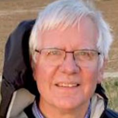 Richard Jozsa, a smiling man with white hair and glasses, He is at the beach and wearing a weatherproof jacket.