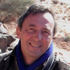 Robert Foley, a smiling man with brown hair wearing a brown jacket and a purple scarf. He is outdoors in a sunny, mountainous region