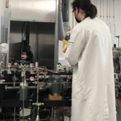 Ross Findlay is facing away from the camera. He is wearing a long white lab coat and is working in front of come machinery. He has long lark hair and is wearing a face mask and gloves
