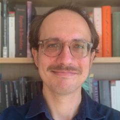 Ryan Heuser, a smiling man with short brown hair, a moustache and glasses. He is wearing a blue shirt and sitting in front of a book shelf
