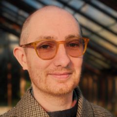 Samuel Moore, a smiling man with a shaved head and yellow glasses. He is wearing a tweed tailored jacket and is standing in building with large windows