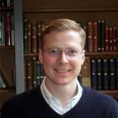 Sebastian Eves van-den Akker, a smiling man with short blonde hair and glasses. He is wearing a navy blue sweater and is standing in front of a book shelf