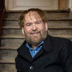 Simon Goldhill, a smiling man with brown hair and a beard wearing a blue shirt and a black jacket. He is sitting on a wooden staircase
