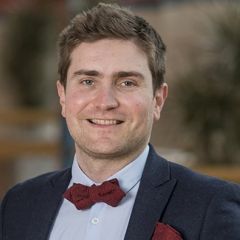 Thomas Roulet, a smiling man with short brown hair, wearing a blue suit with a maroon bow tie