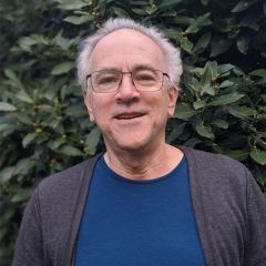 Tim Griffin, a smiling man with short grey hair and glasses wearing a blue t-shirt and a grey cardigan. He is outdoors standing in front of a bush.
