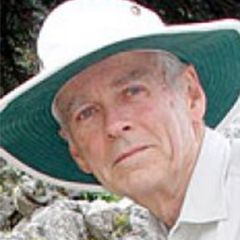 A smiling man with grey hair wearing a wide brimmed white and green sun hat and a white shirt. He is standing in a rocky area