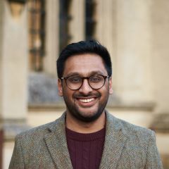 Umang Bhatt, a smiling man with dark hair, a short beard and glasses. He is wearing a maroon jumper and a grey tweed jacket. He is standing outside a Cambridge College