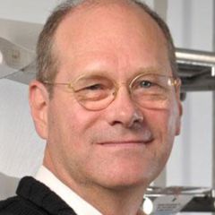 William Baker, a smiling man with short grey hair and glasses wearing a white shirt and a jumper.