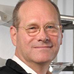 William Baker, a smiling man with short grey hair and glasses wearing a white shirt and a jumper.