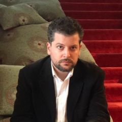 A man in a dark jacket and a white shirt in front of a red staircase
