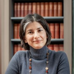 Cryssi Giannitsarou, smiling woman with short dark hair, a grey jumper and a beaded necklace sitting in front of a bookshelf 