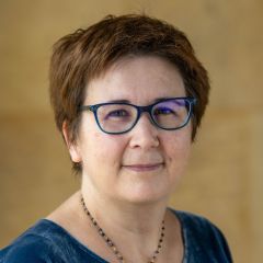 Sophie Turenne, a woman with short auburn hair, blue glasses, a gold necklace and a blue velvet top stands in front of a yellow stone wall