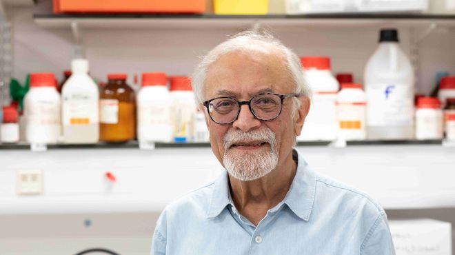 A smiling man with short white hair, a beard and glasses, wearing a pale blue shirt in a lab with bottles on a shelf behind him.