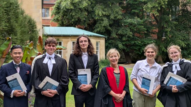 Five people wearing smart academic clothes smile holding a book and a certificate. There is a smiling woman in the middle wearing a black gown with Red detail at the front. They are in a formal garden on a sunny day.